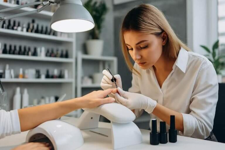 Manichiuristă profesionistă aplicând gel pe unghii într-un salon modern, cu lampă UV și produse de manichiură vizibile.